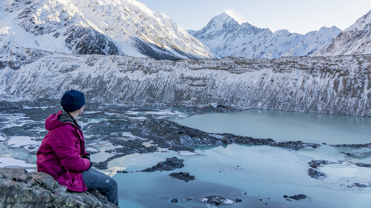 Image of a student sitting down on a large rock looking out to a sea of white, snowy mountains 