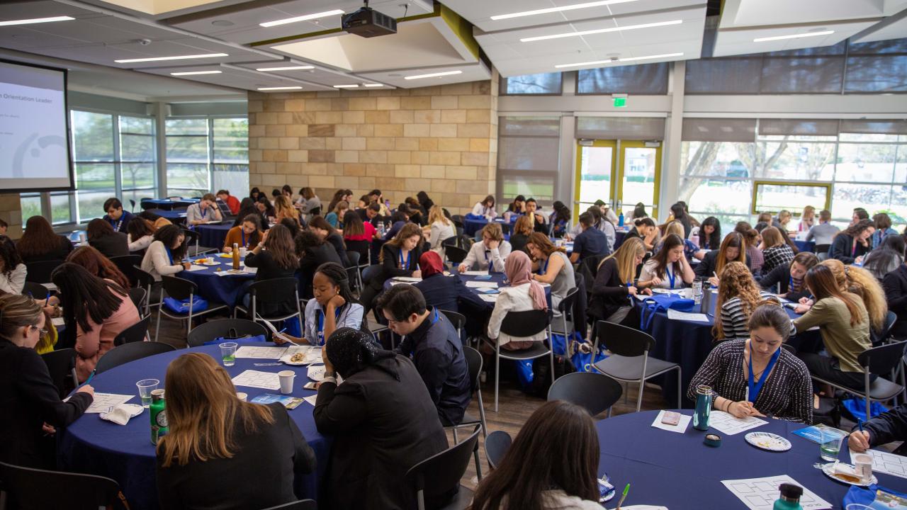 A large group of students sit at round tables in a spacious and bright conference room, engaged in writing and discussing in small groups. The room is filled with natural light from large windows, and a presentation screen is visible in the corner.