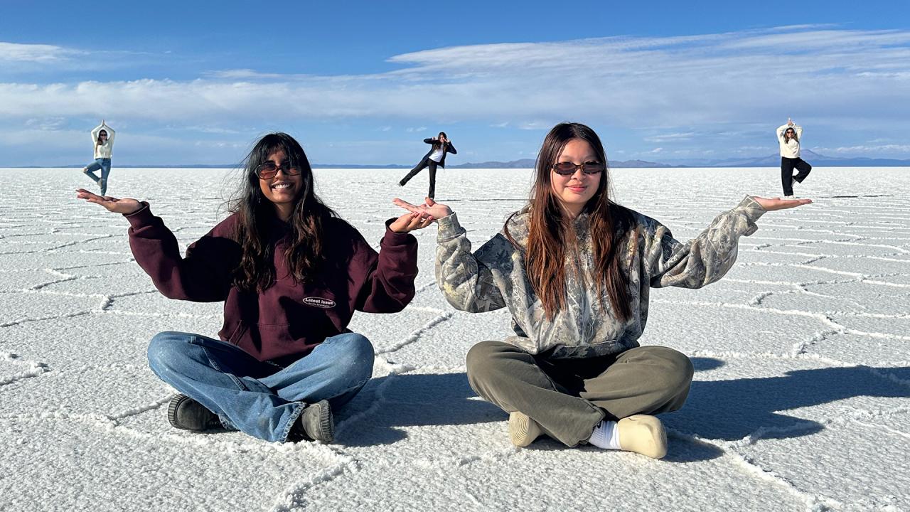 On a sunlit salt flat, two women wearing sunglasses sit on the ground, playfully holding up smaller figures in their hands through forced perspective. The smaller figures in the background stand in yoga poses, with the salt flats stretching out beneath a wide, clear sky.
