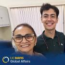 Three people smiling and posing for a selfie indoors, with a backdrop of office furniture and blinds. The group includes a woman wearing glasses, a young man with curly hair, and an older man with gray hair and a mustache. They appear happy and engaged. The image is branded with 'UC Davis Global Affairs' and 'Global Aggies' logos at the bottom corner.