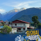 A scenic image of a snow-capped mountain peak at sunrise or sunset, with warm blue light casting over the summit and surrounding village. The UC Davis Global Affairs logo is in the bottom left corner, and the bottom right features the hashtag '#GlobalAggies' with illustrated icons of a bicycle, a globe, and a beaker.