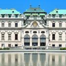 A grand baroque palace with ornate details, the Belvedere in Vienna, Austria, stands prominently with its green copper domes and symmetrical architecture. In front of the palace, a large, reflective pond mirrors the building's façade. The sky is clear and blue, and several people are visible walking along the pathways and garden areas surrounding the palace.