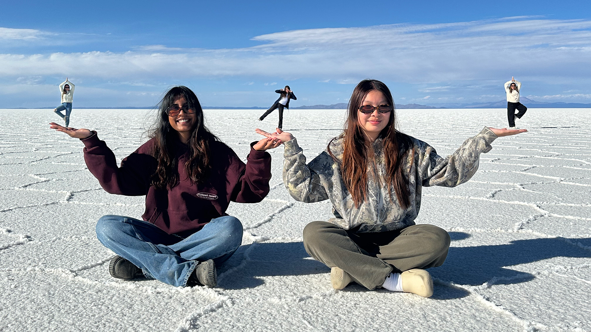 On a sunlit salt flat, two women wearing sunglasses sit on the ground, playfully holding up smaller figures in their hands through forced perspective. The smaller figures in the background stand in yoga poses, with the salt flats stretching out beneath a wide, clear sky.