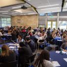 A large group of students sit at round tables in a spacious and bright conference room, engaged in writing and discussing in small groups. The room is filled with natural light from large windows, and a presentation screen is visible in the corner. 