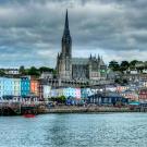 A waterfront view of Cobh, Ireland, featuring St. Colman’s Cathedral with its tall Gothic spire rising above rows of colorful buildings. The town's houses are painted in bright hues of blue, yellow, and pink, contrasting with the overcast sky. A small red boat floats in the foreground on calm water.