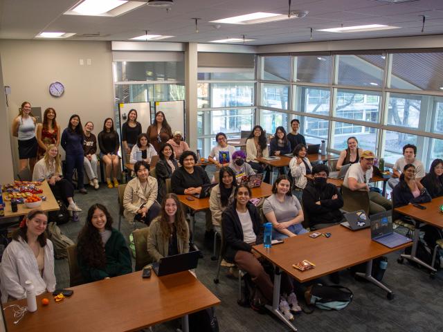 A diverse group of students smiling in a classroom setting with tables and large windows.