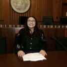 Smiling woman with glasses at wood-paneled council table with papers, photo
