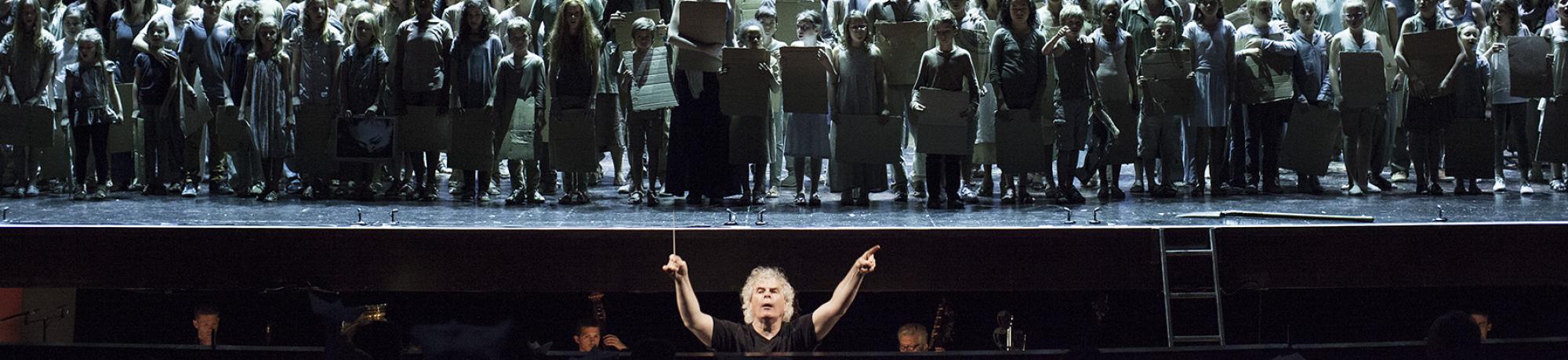A large group of performers on stage with a wall of black-and-white portraits behind them.