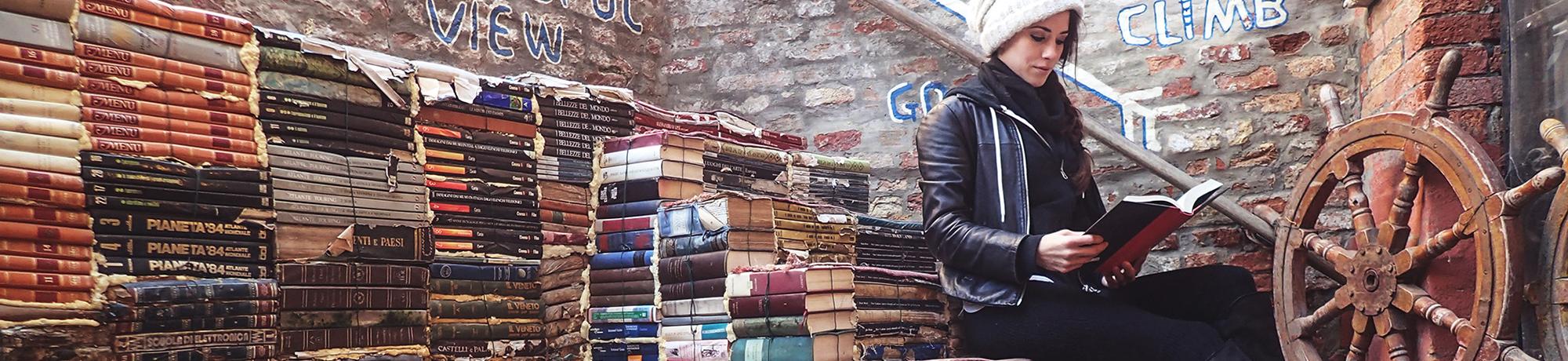 A woman reads a book while sitting on stacks of old books in a cozy, cluttered space.